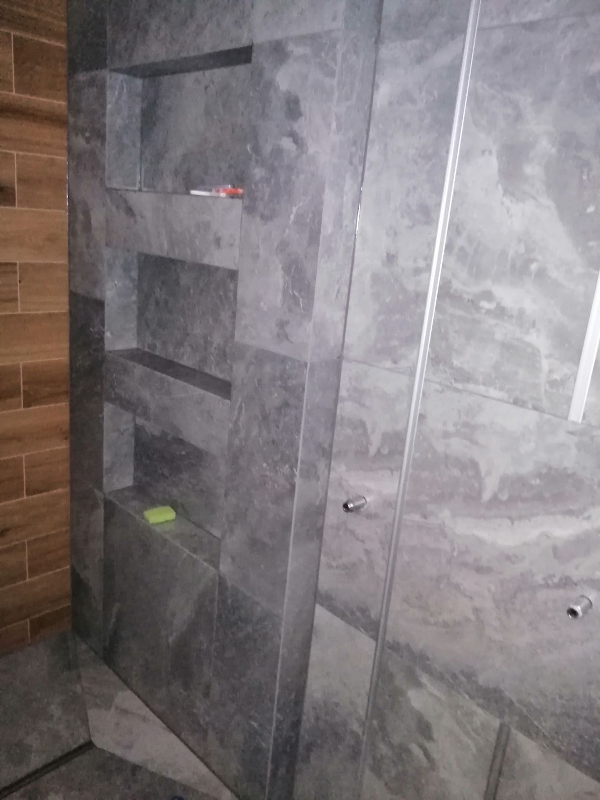 Szara, marmurkowa ściana prysznicowa z wnękami, na jednej z nich leży zielone mydło, obok widoczna drewnopodobna okładzina.