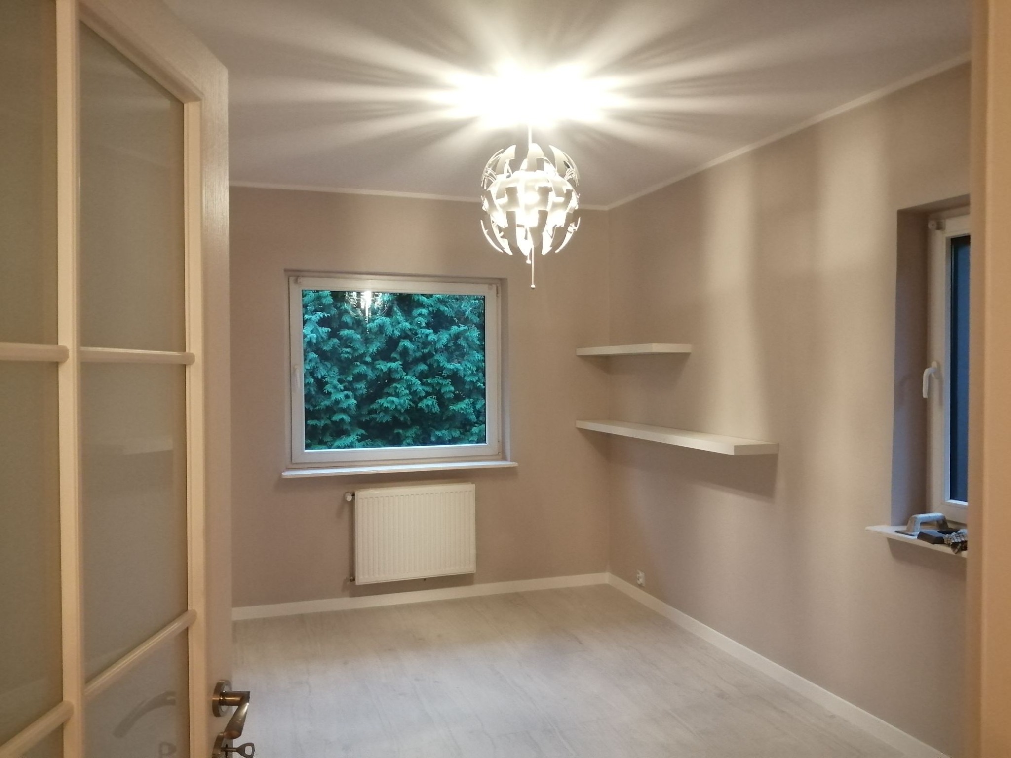 Pomalowane na beżowo wnętrze pokoju z jasną podłogą, oknem z widokiem na zieleń, białym grzejnikiem i designerską lampą sufitową.