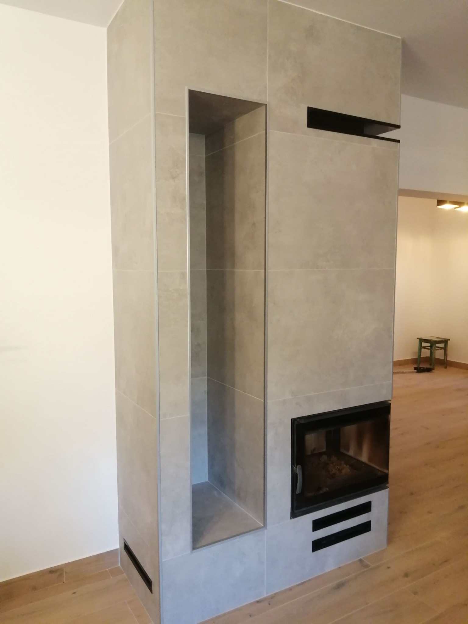 Obudowa kominka wykończona płytkami ceramicznymi w odcieniach szarości, z wnęką na drewno i czarnymi elementami dekoracyjnymi, na tle drewnianej podłogi i fragmentu białej ściany.