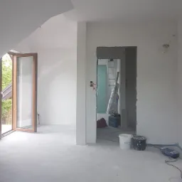 Wnętrze w trakcie remontu: świeżo pomalowane na biało ściany poddasza, otwarte okno, widoczne narzędzia i wiaderka po farbie, przygotowane do dalszych prac wykończeniowych.