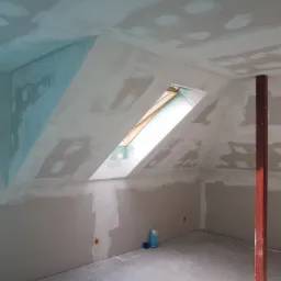 Poddasze w trakcie prac wykończeniowych: ściany i sufit pokryte częściowo gładzią, widoczne łączenia płyt kartonowo-gipsowych, okno dachowe w skosie, metalowy słup konstrukcyjny i butelka z płynem...
