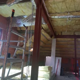 Ocieplone poddasze z widoczną konstrukcją stalową, wełną mineralną i folią aluminiową, drabina aluminiowa i stos płyt gipsowo-kartonowych na podłodze.