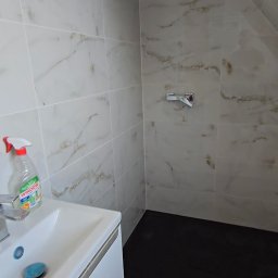 TOM-KAR - Wykończona łazienka z marmurowymi płytkami na ścianach, czarną podłogą i białą szafką pod umywalką. Widoczny kran i butelka płynu do mycia szyb.