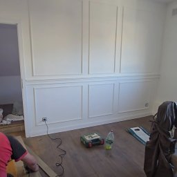 TOM-KAR Usługi Kosztorysowo-Budowlane Karina TOMCZYK - Montaż paneli podłogowych w pokoju z dekoracyjną sztukaterią ścienną. Widoczny fragment osoby przycinającej panel, narzędzia i butelka wody.