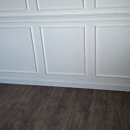 TOM-KAR Usługi Kosztorysowo-Budowlane Karina TOMCZYK - Biała ściana z dekoracyjnymi panelami i ciemna podłoga z paneli. Widok na fragment wykończonego wnętrza z wyraźnym kontrastem kolorystycznym.