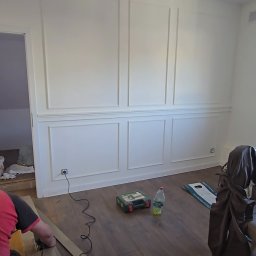 TOM-KAR Usługi Kosztorysowo-Budowlane Karina TOMCZYK - Montaż paneli podłogowych w pokoju z dekoracyjną sztukaterią ścienną. Widoczny fragment osoby przycinającej panel, narzędzia i butelka wody.