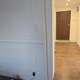 TOM-KAR Usługi Kosztorysowo-Budowlane Karina TOMCZYK - Wykończone wnętrze: biała ściana z dekoracyjnymi panelami, brązowe panele podłogowe, widok na korytarz z drewnianymi drzwiami. W tle widoczne narzędzia.