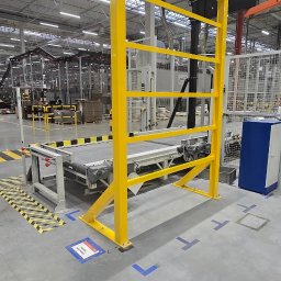 TOM-KAR Usługi Kosztorysowo-Budowlane Karina TOMCZYK - Żółta, metalowa konstrukcja ochronna przy linii produkcyjnej w hali. Widoczne oznaczenia BHP na podłodze i elementy linii produkcyjnej w tle.