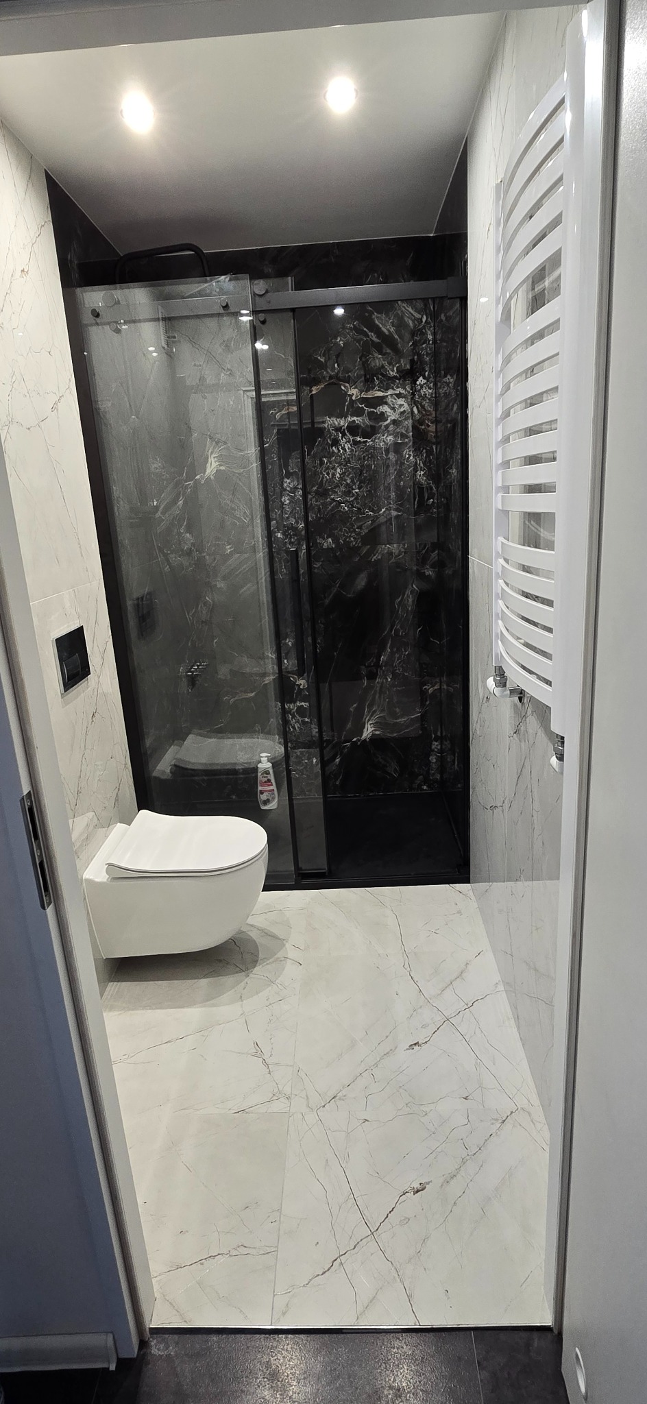 Nowoczesna łazienka po remoncie: białe marmurowe płytki, kabina prysznicowa z czarnymi ramami, podwieszana toaleta i grzejnik drabinkowy. Minimalistyczny design.