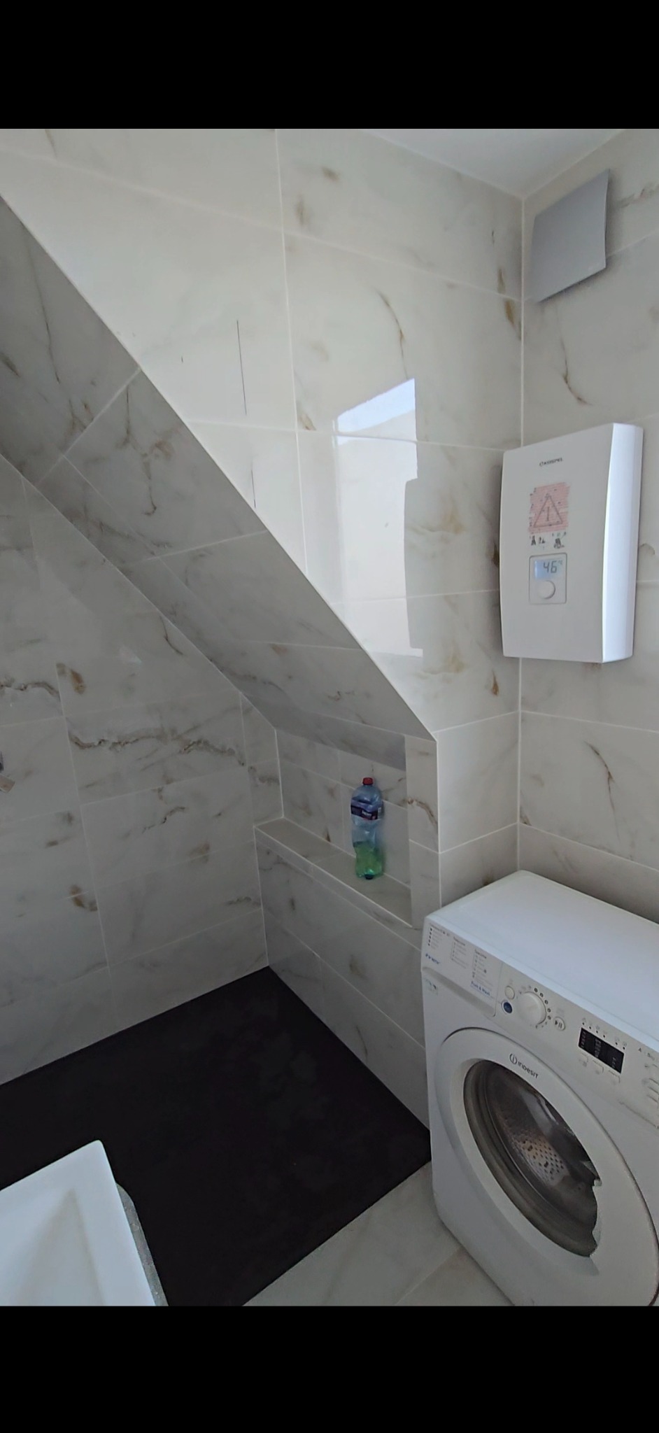 Wykończona łazienka na poddaszu z marmurowymi płytkami, białą pralką i czarną podłogą. Wnęka w ścianie z butelką. Widoczny wentylator i grzejnik.