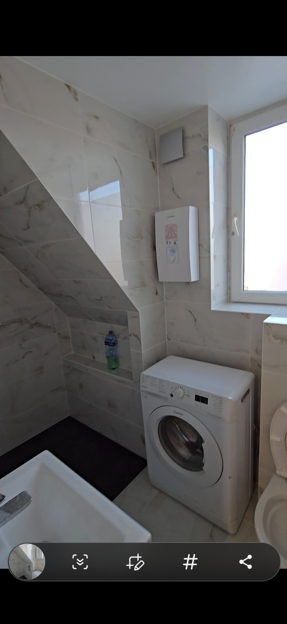 Wykończona łazienka na poddaszu z marmurowymi płytkami, białą pralką i bojlerem. Umywalka z baterią i toaleta widoczne w kadrze. Okno z białymi ramami.