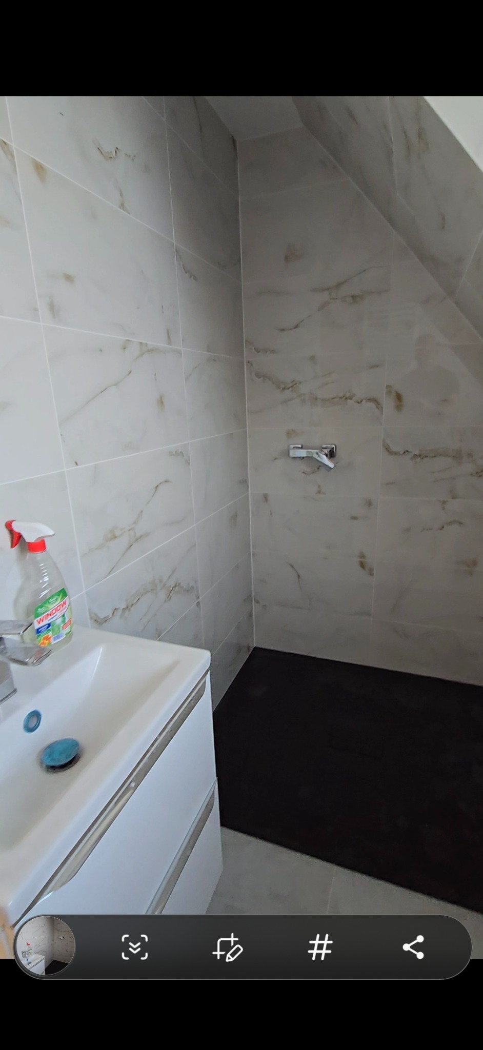 Wykończona łazienka z marmurowymi płytkami na ścianach, czarną podłogą i białą szafką pod umywalką. Widoczny kran i butelka płynu do mycia szyb.