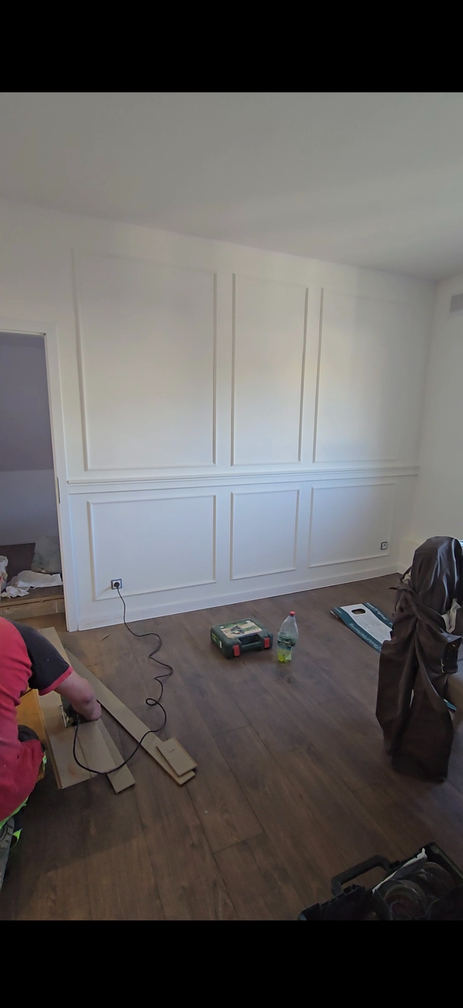 Montaż paneli podłogowych w pokoju z dekoracyjną sztukaterią ścienną. Widoczny fragment osoby przycinającej panel, narzędzia i butelka wody.