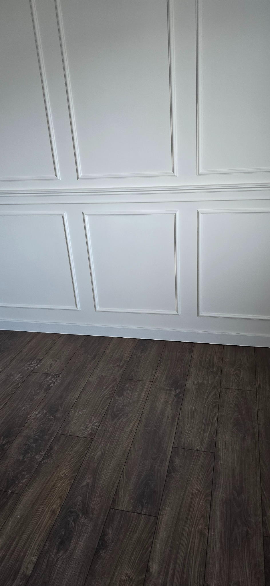 Biała ściana z dekoracyjnymi panelami i ciemna podłoga z paneli. Widok na fragment wykończonego wnętrza z wyraźnym kontrastem kolorystycznym.