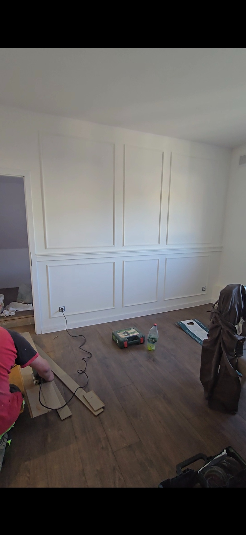 Montaż paneli podłogowych w pokoju z dekoracyjną sztukaterią ścienną. Widoczny fragment osoby przycinającej panel, narzędzia i butelka wody.