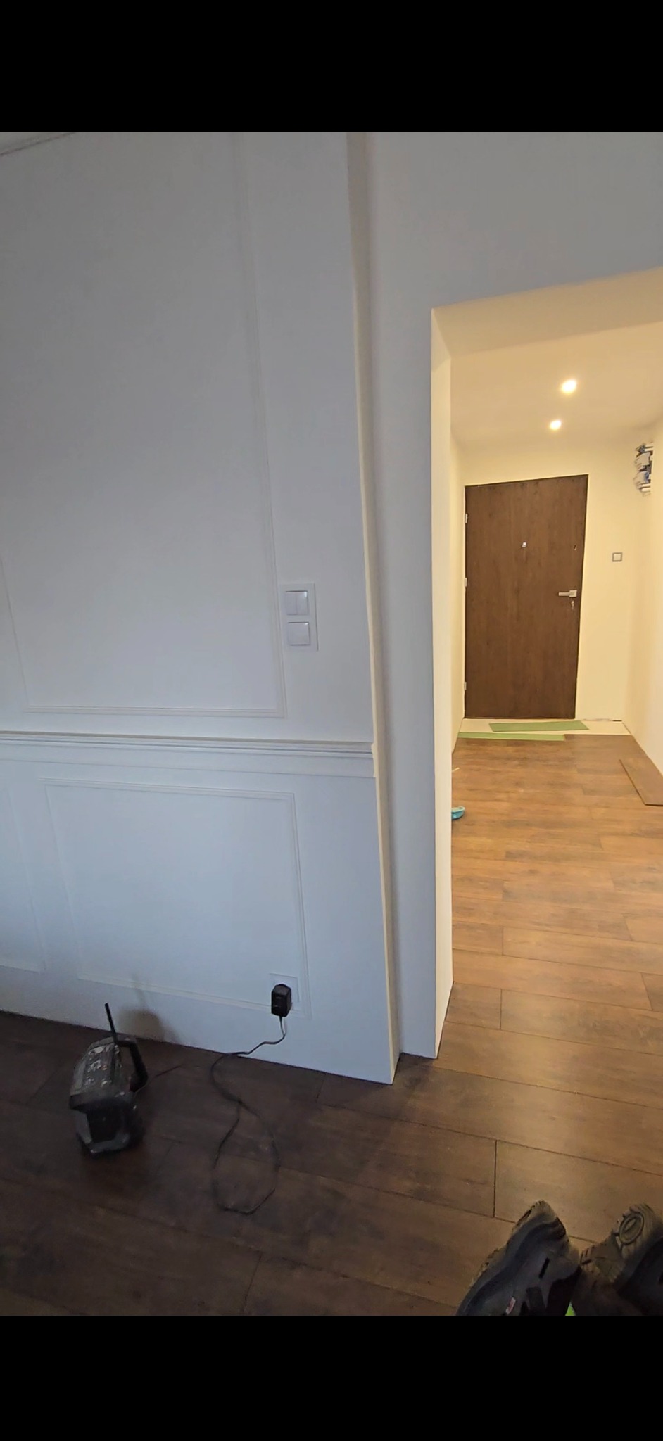 Wykończone wnętrze: biała ściana z dekoracyjnymi panelami, brązowe panele podłogowe, widok na korytarz z drewnianymi drzwiami. W tle widoczne narzędzia.