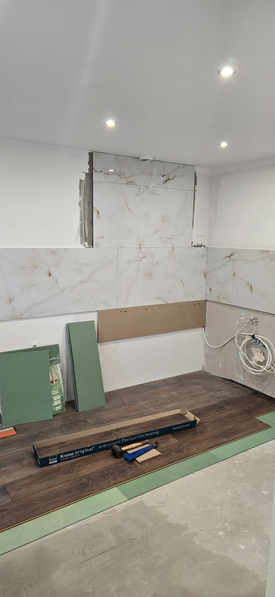 Wnętrze w trakcie remontu: częściowo ułożone panele podłogowe, płytki ścienne z marmurowym wzorem, narzędzia i materiały budowlane. Widoczne niedokończone prace.