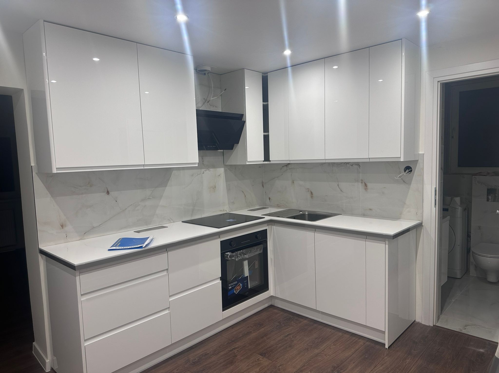 Nowoczesna, biała kuchnia z połyskiem, marmurowy backsplash, czarny okap i płyta indukcyjna. Widok na łazienkę z pralką. Drewniana podłoga.