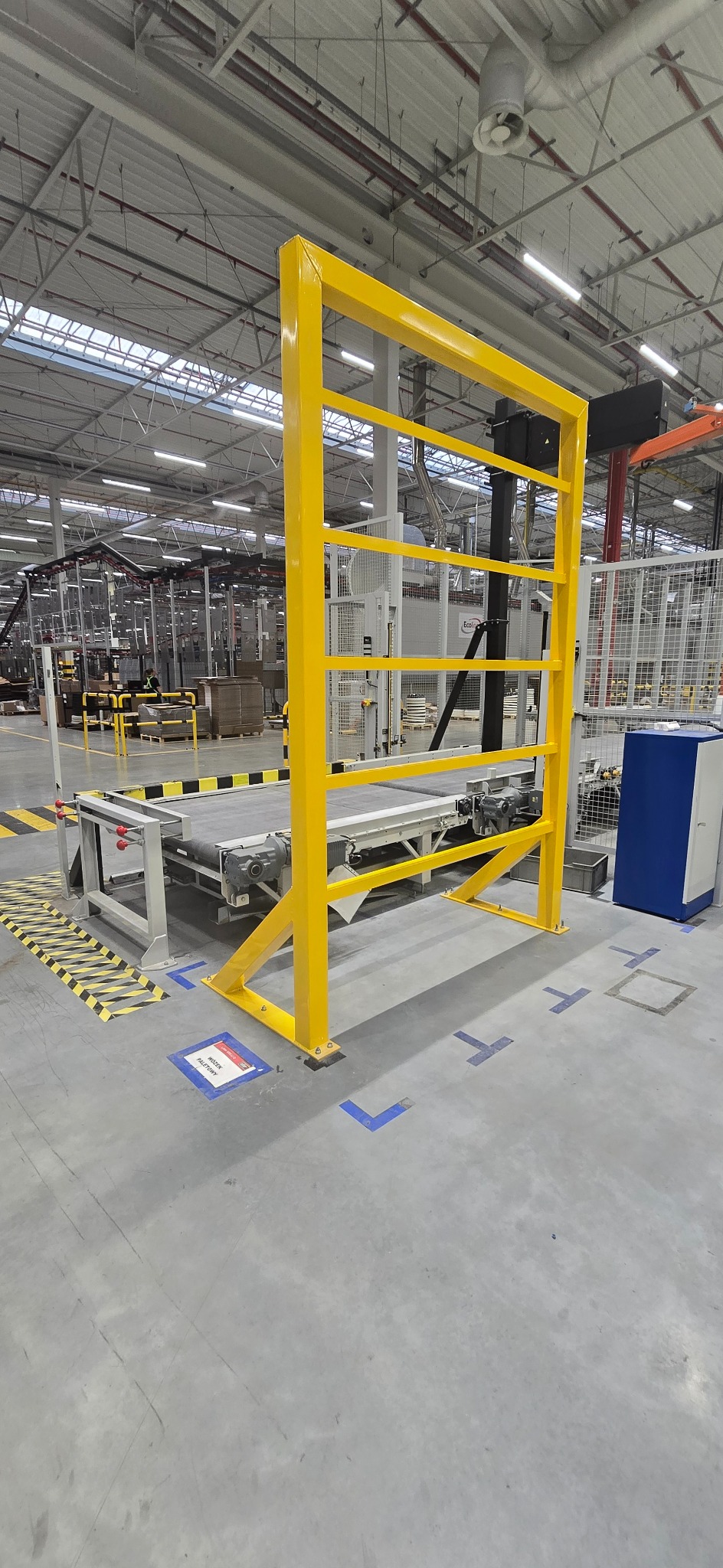 Żółta, metalowa konstrukcja ochronna przy linii produkcyjnej w hali. Widoczne oznaczenia BHP na podłodze i elementy linii produkcyjnej w tle.