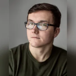 Portret młodego mężczyzny w okularach z czarnymi oprawkami, ubrany w zieloną koszulkę, na jednolitym szarym tle.