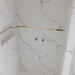 Wykończona kabina prysznicowa z białymi płytkami imitującymi marmur, złotymi listwami dekoracyjnymi i punktowymi przyłączami wodnymi.