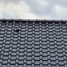 Błyszczące, ciemnoszare dachówki ceramiczne ułożone na dachu, widok z dołu na tle pochmurnego nieba. W centralnej części dachu widoczne pojedyncze uszkodzenie lub brak dachówki.