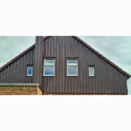 Dom z elewacją z brązowej blachy trapezowej, widoczne okna z białymi ramami i ceglana podmurówka.