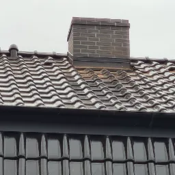 Widok z góry na brązowy dach z dachówki ceramicznej z kominem z cegły klinkierowej, widoczne orynnowanie i fragment ściany budynku pokrytej okładziną imitującą dachówkę.