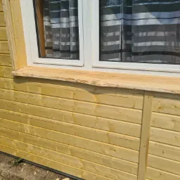 Drewniana elewacja budynku z oknem, surowa deska parapetu z widoczną korą, szare zasłony w paski w oknie.