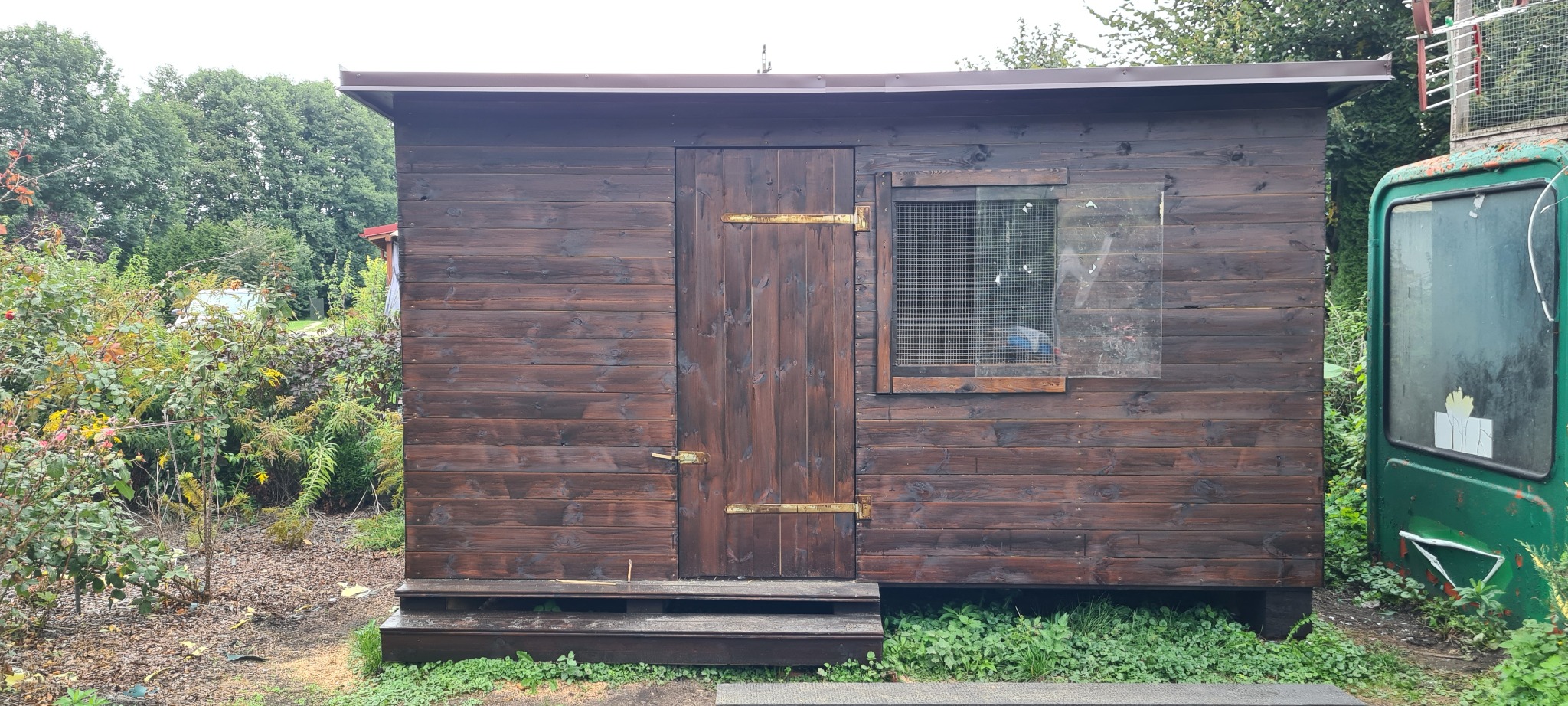 Drewniana konstrukcja typu ptaszarnia z brązowymi deskami, małym oknem z siatką i częściowo przyklejoną folią, oraz brązowym dachem, otoczona zielenią.