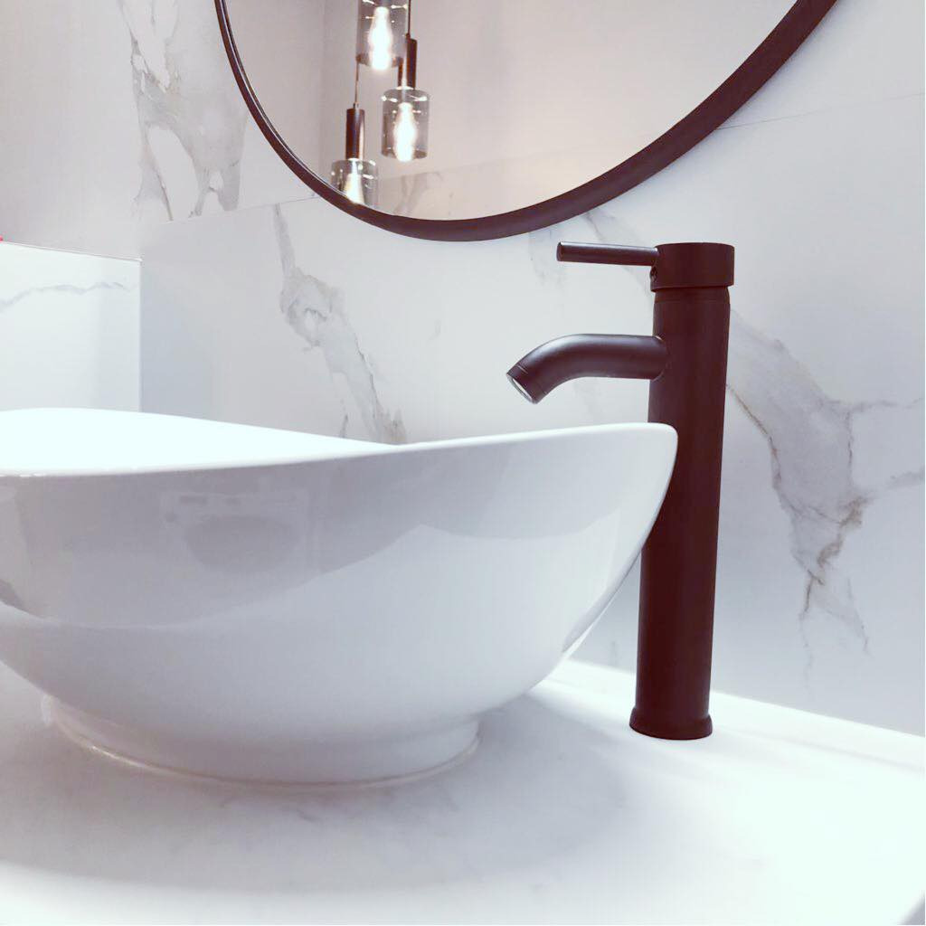 Biała, okrągła umywalka nablatowa z czarną, minimalistyczną baterią na tle marmurowej ściany i okrągłego lustra z widocznymi wiszącymi lampami.