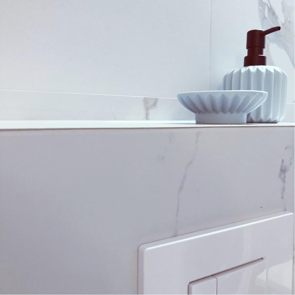 Biała łazienka z marmurowym wykończeniem, minimalistyczna umywalka z dozownikiem mydła i mydelniczką o falistym wzorze, widoczny panel spłuczki.