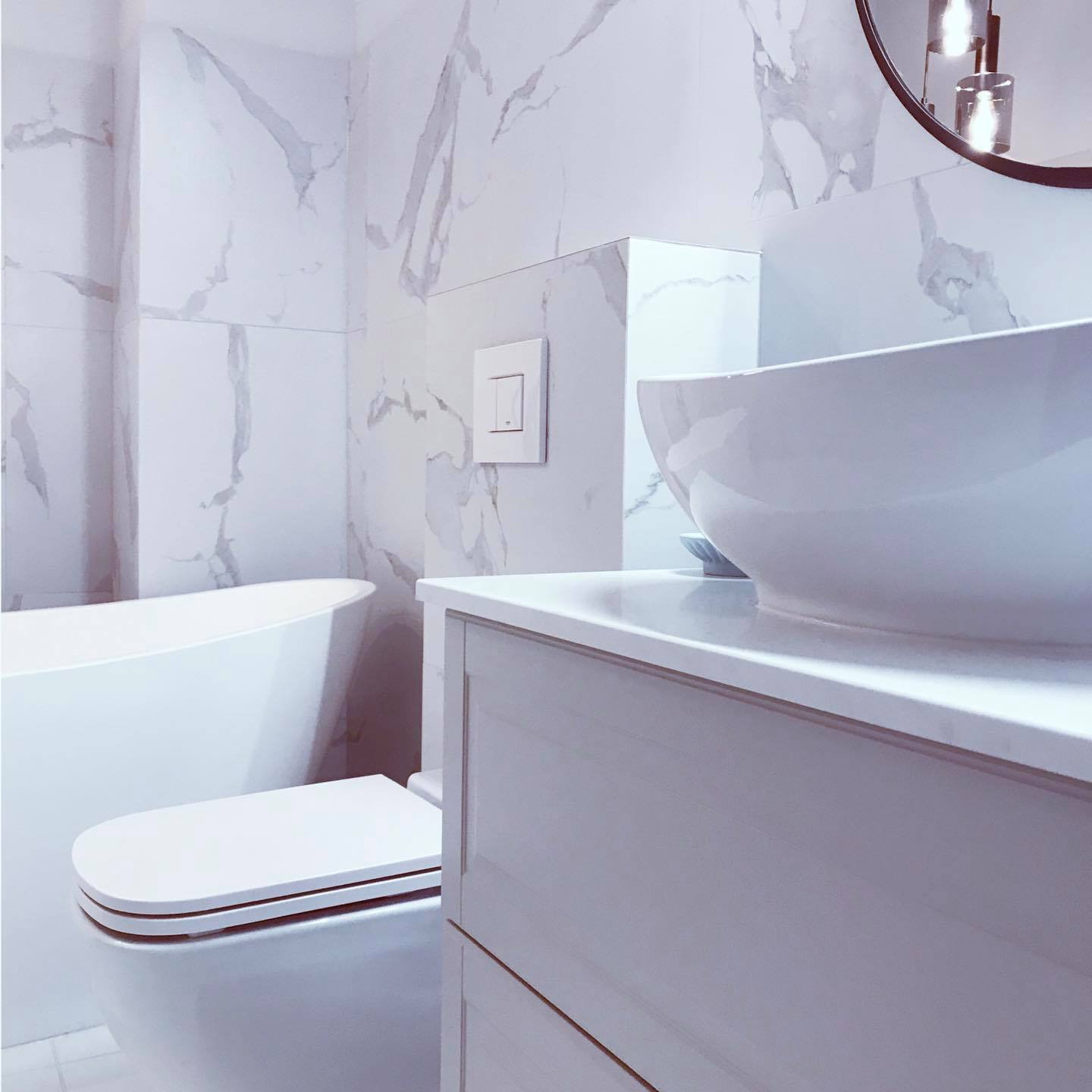 Nowoczesna, jasna łazienka z marmurowymi płytkami, wolnostojącą białą wanną, podwieszaną toaletą i okrągłym lustrem z oświetleniem punktowym.