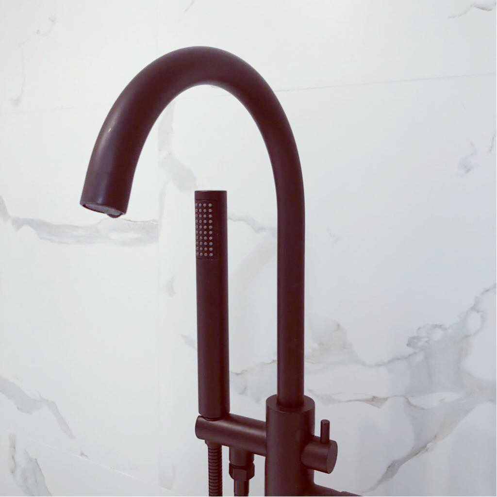 Matowa, czarna bateria wannowa wolnostojąca z słuchawką prysznicową na tle marmurowej ściany.