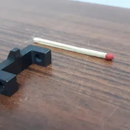 Czarny, metalowy element o nieregularnym kształcie z dwoma otworami, leżący na drewnianym blacie obok zapałki dla porównania skali.