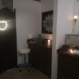 Centrum Regeneracji - gabinet masażu z elementami rehabilitacji Maciejewska Małgorzata - Wnętrze gabinetu z dekoracyjnym parawanem, umywalką na szafce, okrągłym stołkiem i podgrzewaczem do ręczników z ozdobnymi świecami i ręcznikami przewiązanymi wstążkami.