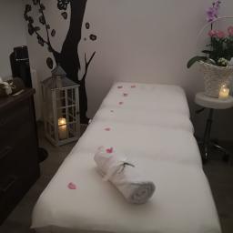 Centrum Regeneracji - gabinet masażu z elementami rehabilitacji Maciejewska Małgorzata - Przygotowane stanowisko do masażu z białym prześcieradłem, ręcznikiem z kokardą, płatkami róż, lampionem ze świecą i dekoracją drzewa na ścianie w tle.