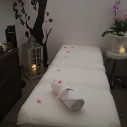 Przygotowane stanowisko do masażu z białym prześcieradłem, ręcznikiem z kokardą, płatkami róż, lampionem ze świecą i dekoracją drzewa na ścianie w tle.