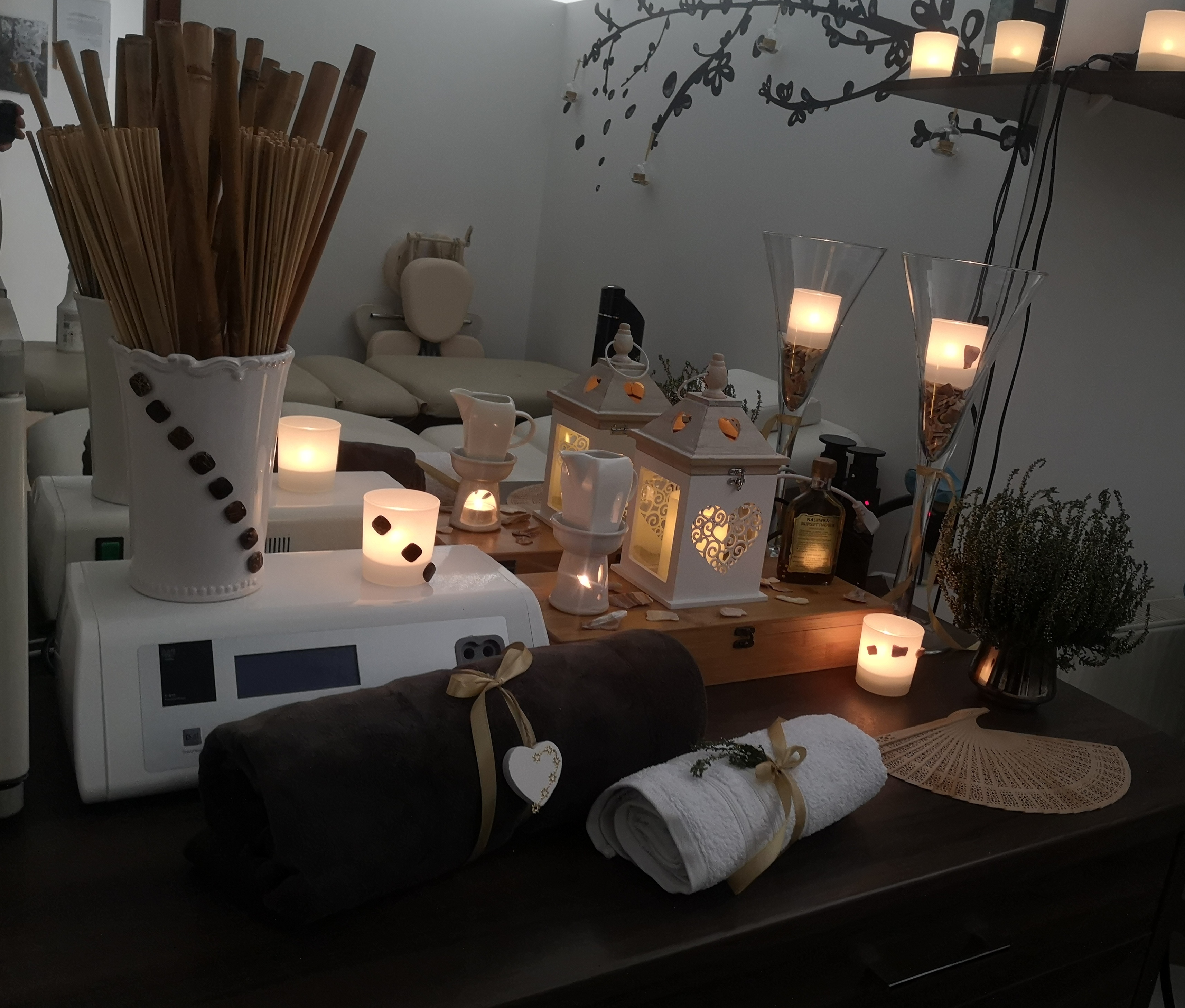 Przytulne wnętrze gabinetu: bambusowe kije w ozdobnym wazonie, zwinięte ręczniki z kokardami, lampiony i świece tworzą relaksującą atmosferę, w tle widoczny stół do zabiegów.