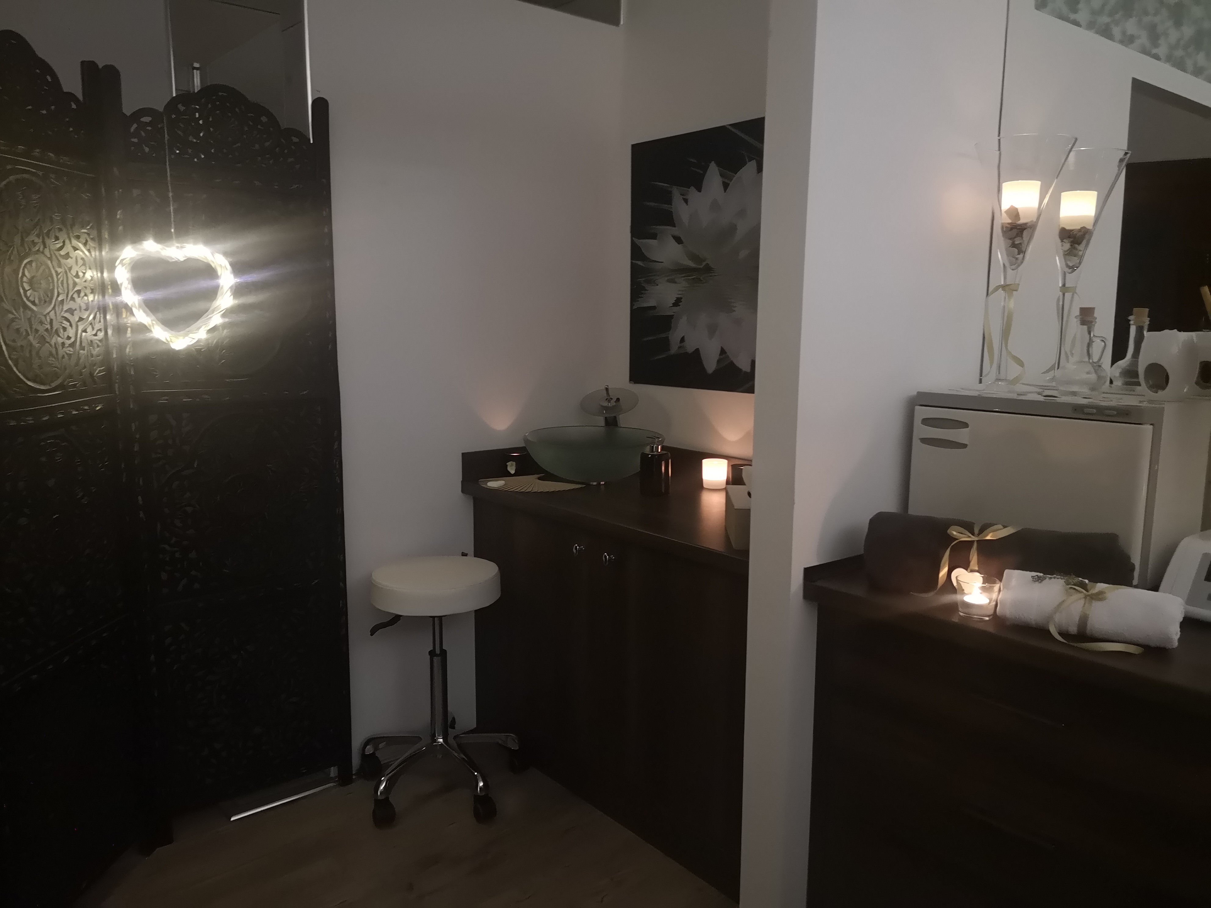 Wnętrze gabinetu z dekoracyjnym parawanem, umywalką na szafce, okrągłym stołkiem i podgrzewaczem do ręczników z ozdobnymi świecami i ręcznikami przewiązanymi wstążkami.