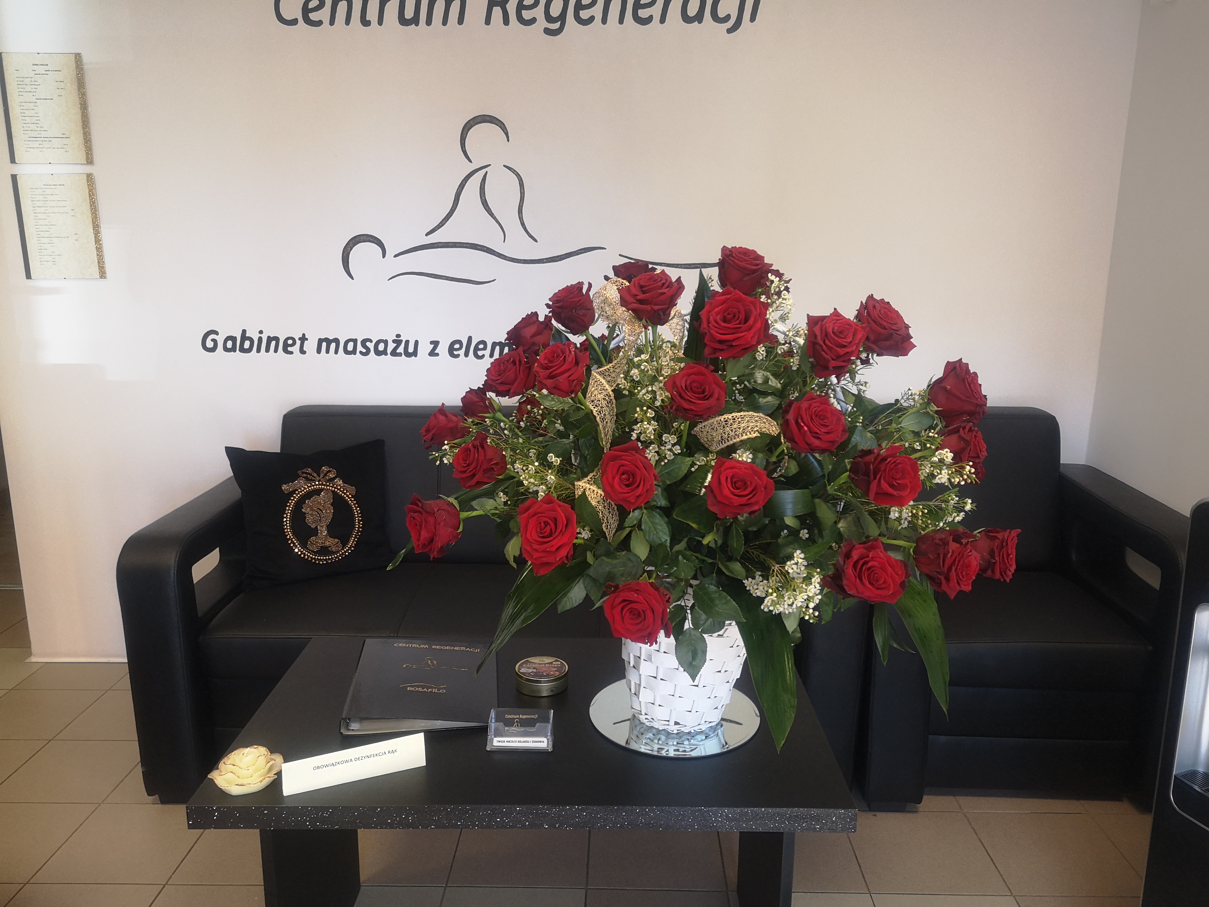 Wnętrze gabinetu masażu z dekoracyjnym bukietem czerwonych róż w białym koszyku, czarną sofą z poduszką z motywem korony, stolikiem z informacją o dezynfekcji rąk i logo 'Centrum Regeneracji'.