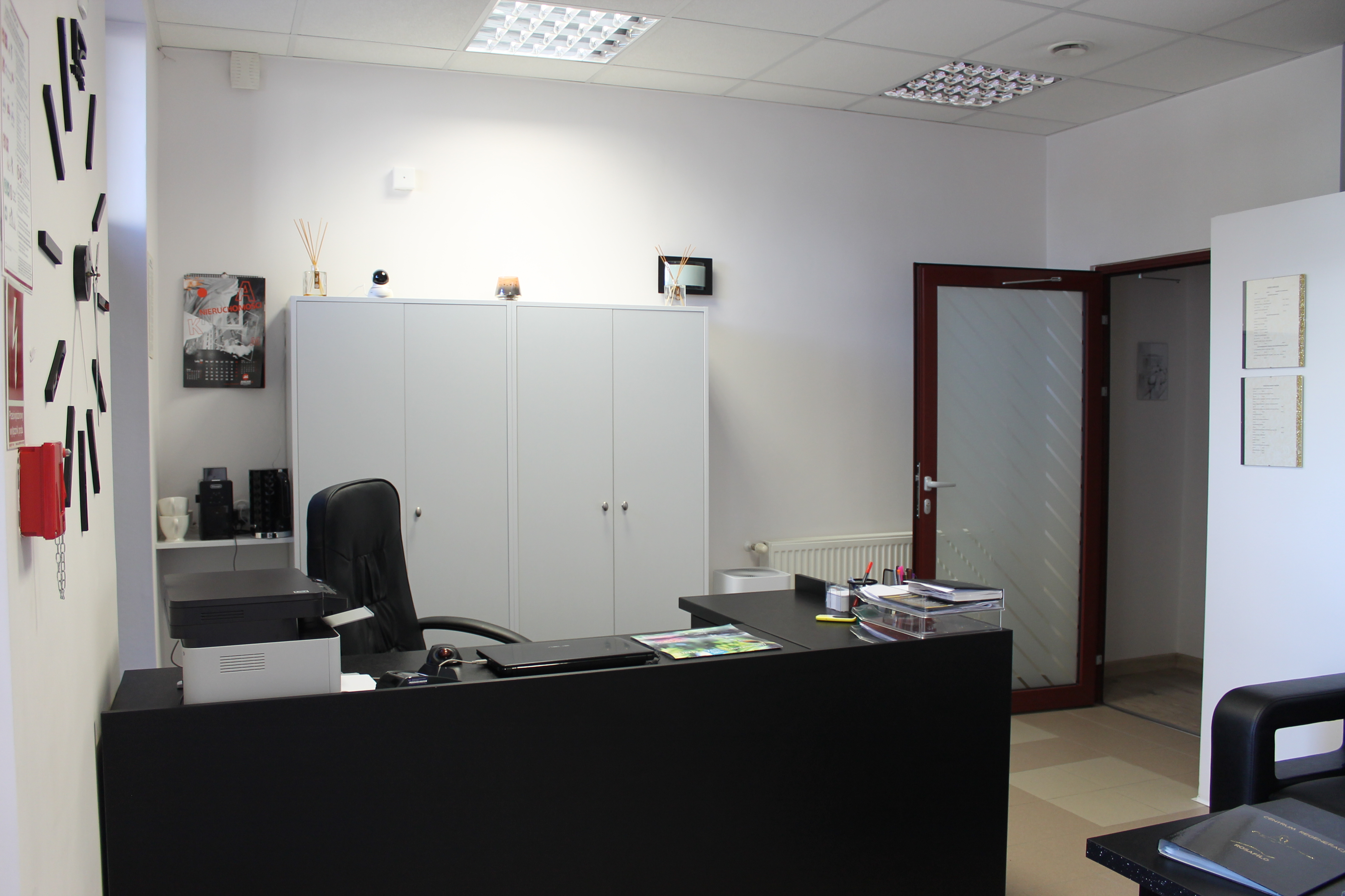 Nowoczesne wnętrze gabinetu z czarnym biurkiem, krzesłem, szafą i elementami dekoracyjnymi, takimi jak dyfuzor zapachowy, kalendarz i minimalistyczny zegar ścienny.