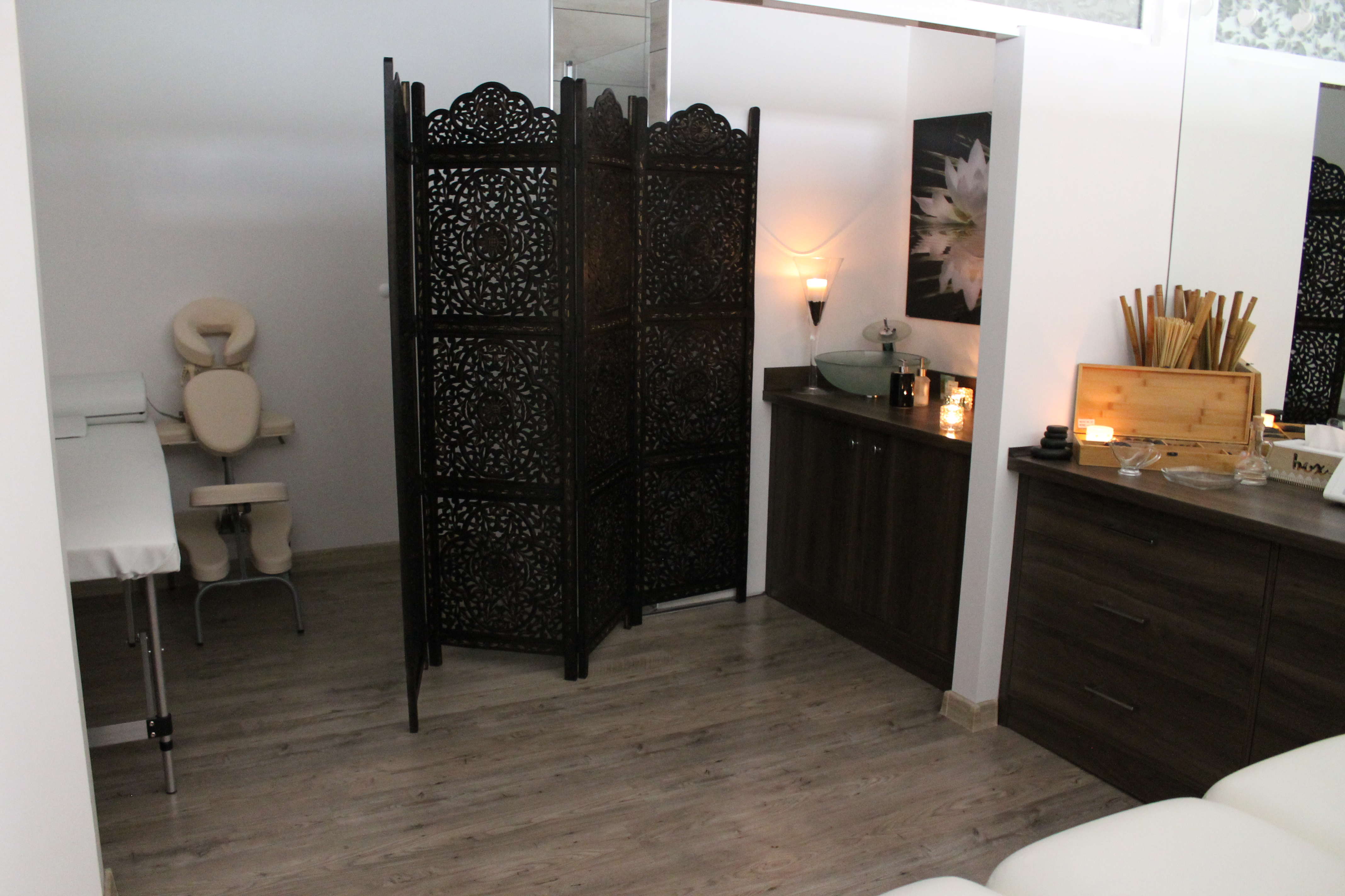 Jasne wnętrze gabinetu z parawanem z ciemnego drewna, stolikiem z umywalką i świecami, oraz leżanką do zabiegów z zagłówkiem.