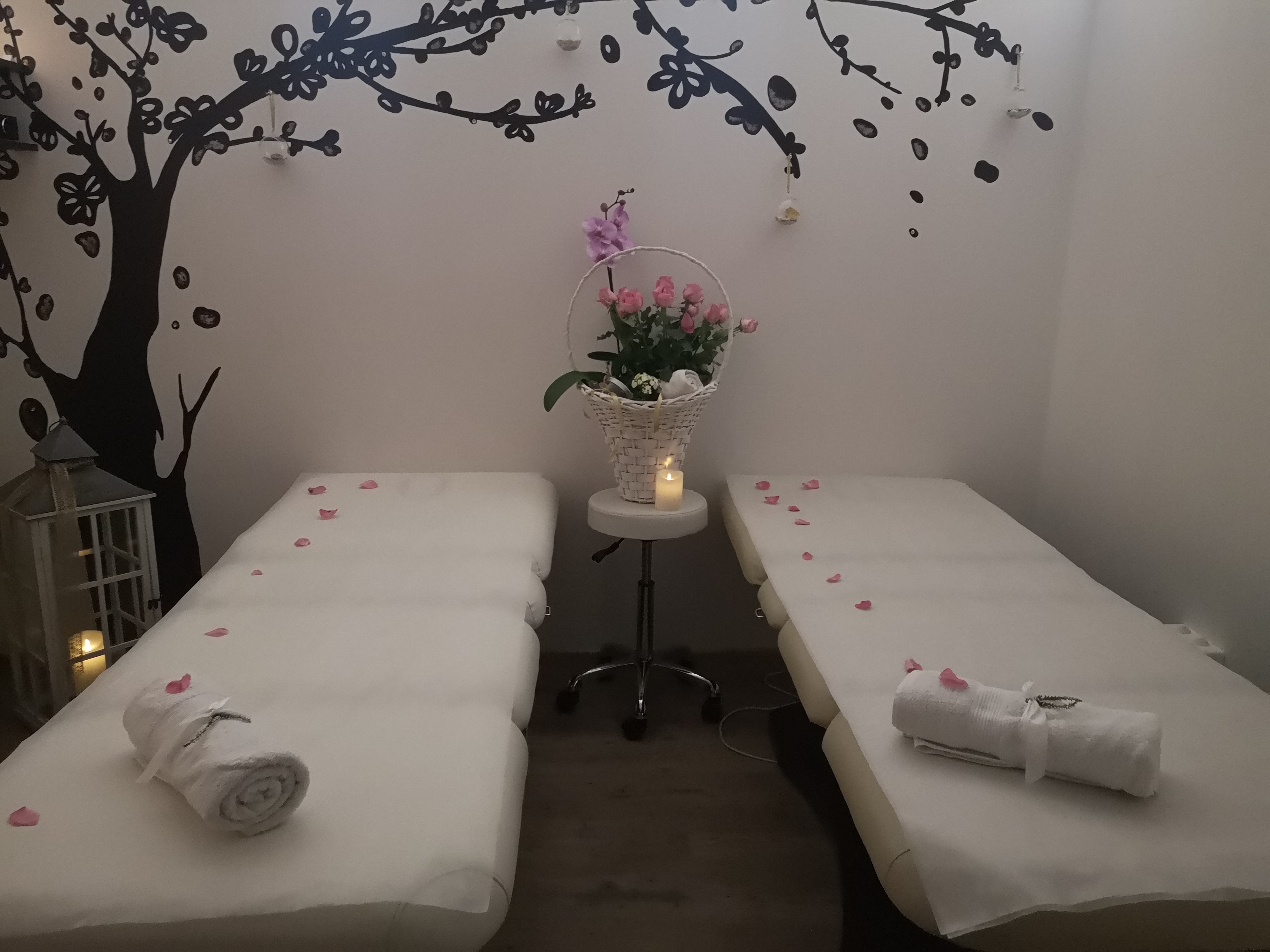 Przygotowane stanowiska zabiegowe z białymi ręcznikami, płatkami róż i dekoracją ścienną w kształcie drzewa, oświetlone świecą na stoliku.