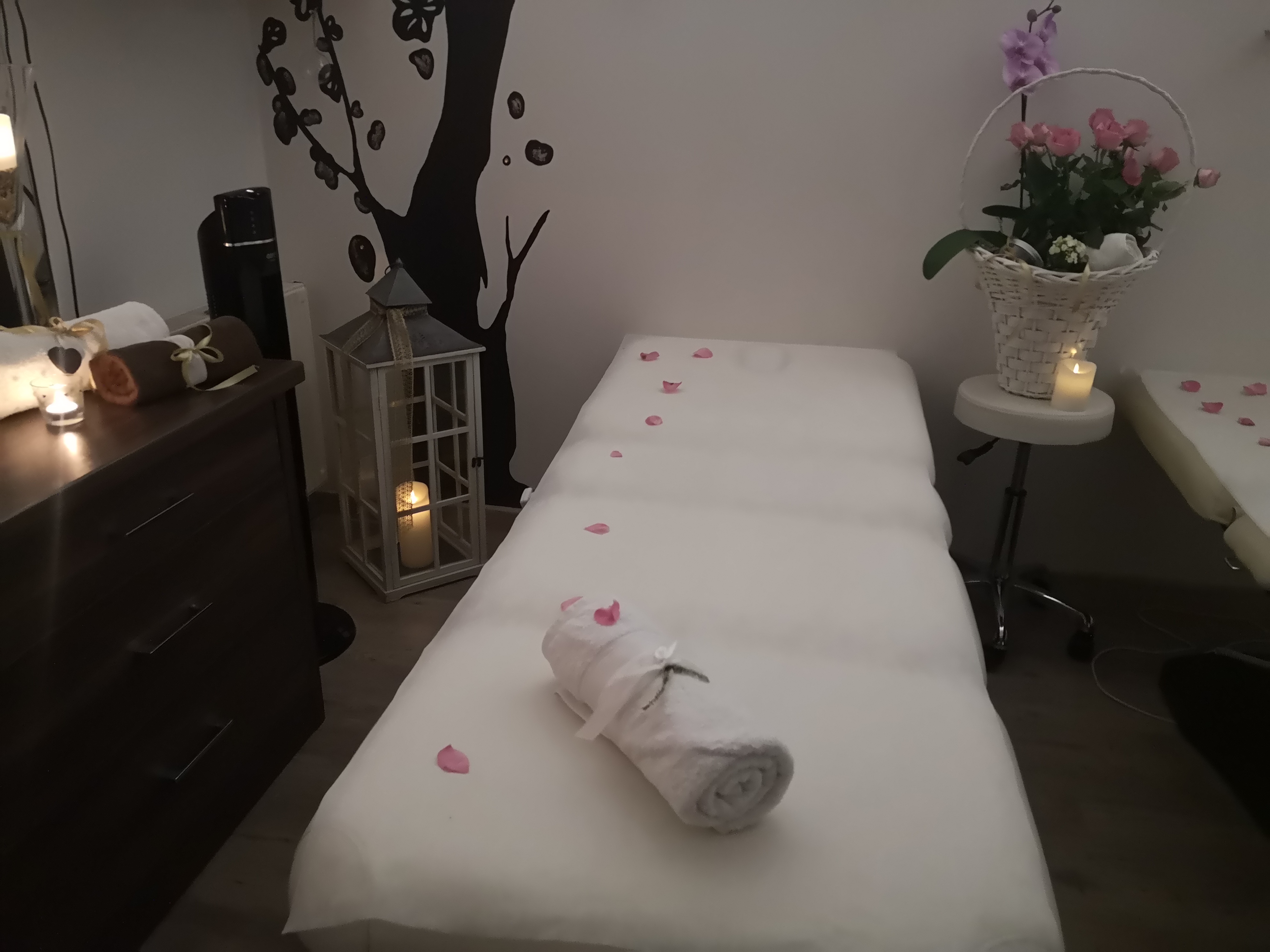 Przygotowane stanowisko do masażu z białym prześcieradłem, ręcznikiem z kokardą, płatkami róż, lampionem ze świecą i dekoracją drzewa na ścianie w tle.