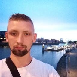 Kowalski De-lux - Mężczyzna z brodą robiący selfie na tle rzeki z budynkami i oświetlonym nabrzeżem o zmierzchu.