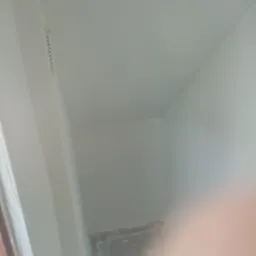 Świeżo pomalowany róg pomieszczenia z widoczną listwą karniszową i folią ochronną na podłodze, ujęcie z bliska z częściowo zasłoniętym obiektywem.