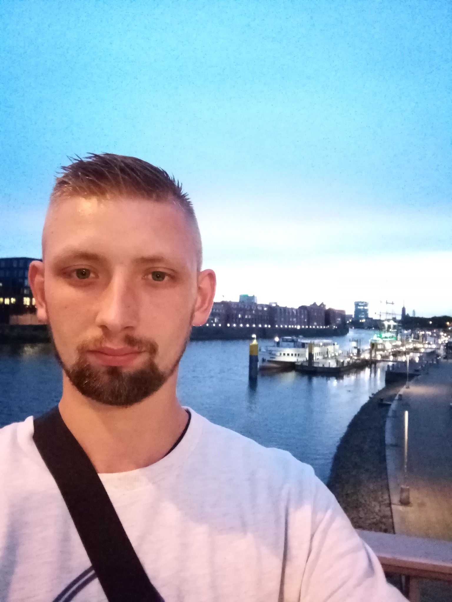 Mężczyzna z brodą robiący selfie na tle rzeki z budynkami i oświetlonym nabrzeżem o zmierzchu.