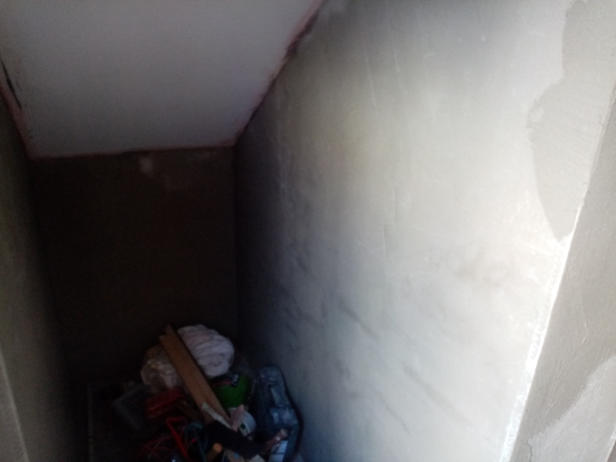 Niedokończone wnętrze ze świeżo otynkowaną ścianą w rogu pomieszczenia, widoczne narzędzia i materiały budowlane w ciemnym kącie.