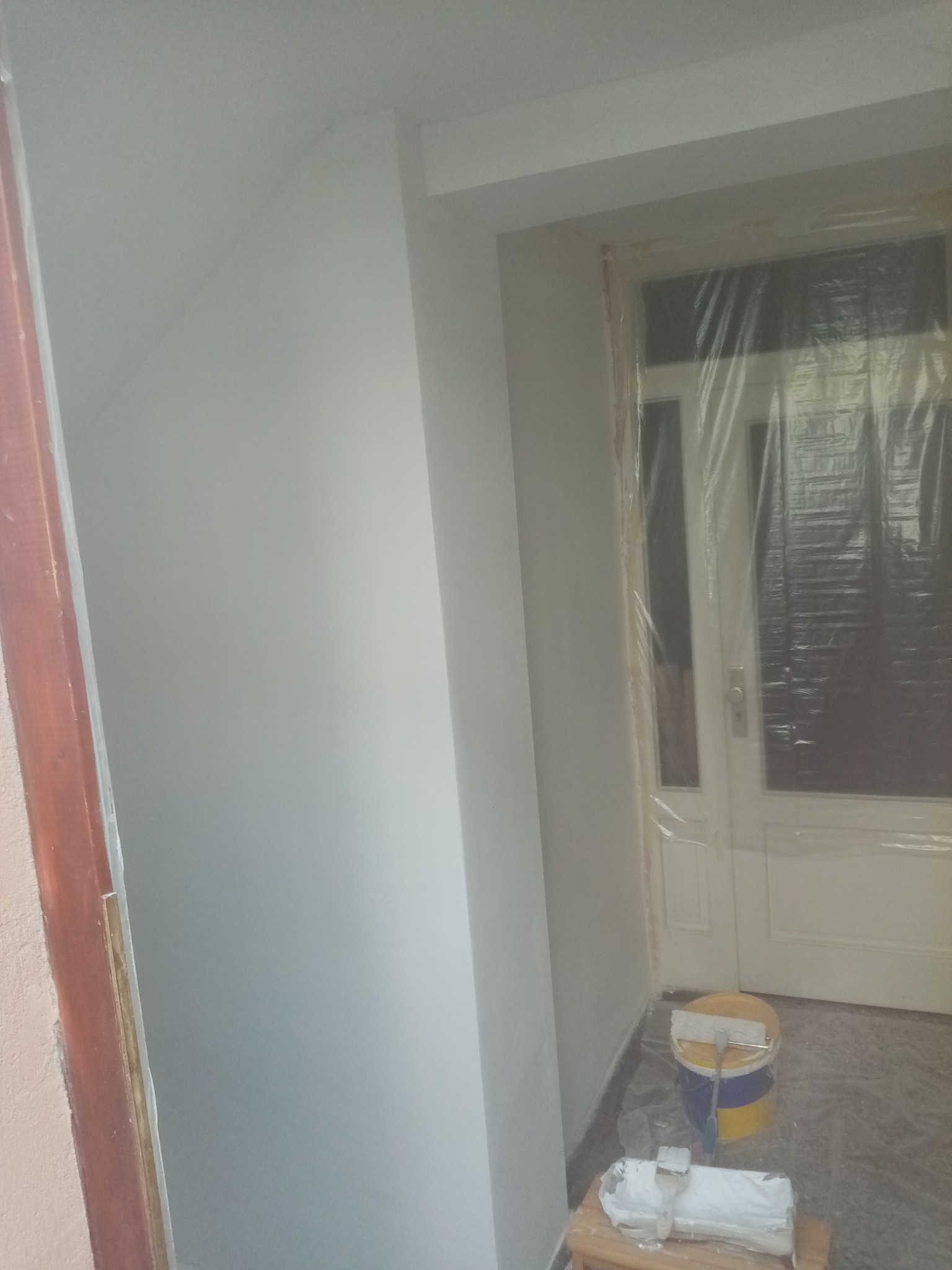 Świeżo pomalowane na biało wnętrze z zabezpieczonymi folią drzwiami, widoczne wiadro z farbą i wałek malarski na podłodze oraz pędzel na małym stołku.